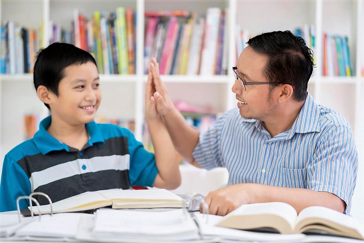 A father and his young son celebrate with a high-five gesture, both wearing big smiles, as they sit at a table with papers and books scattered around them, suggesting they have been working on homework together.