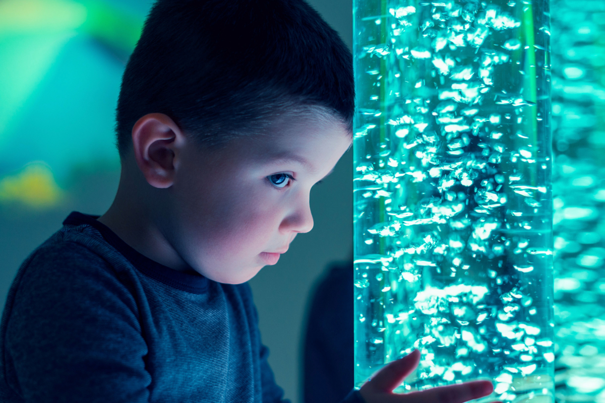 A child interacting with a coloured lights bubble tube lamp in a sensory-stimulating room.