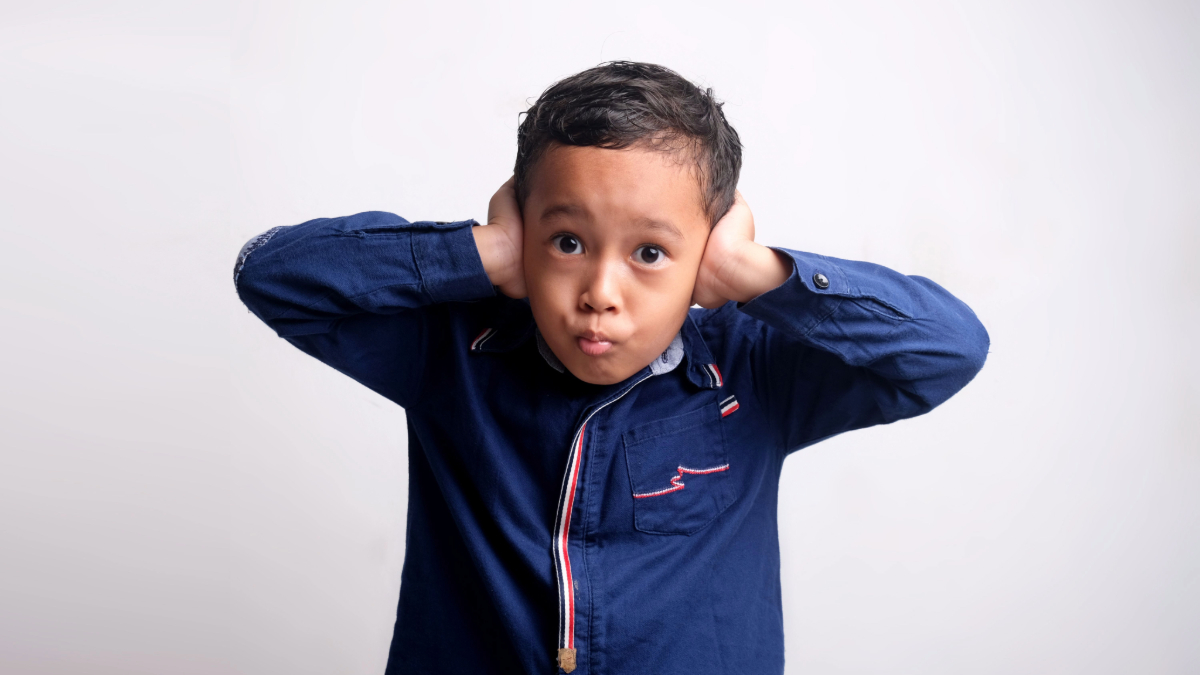A young Asian boy of five to six years old, with an unpleasant look, covers his ears.