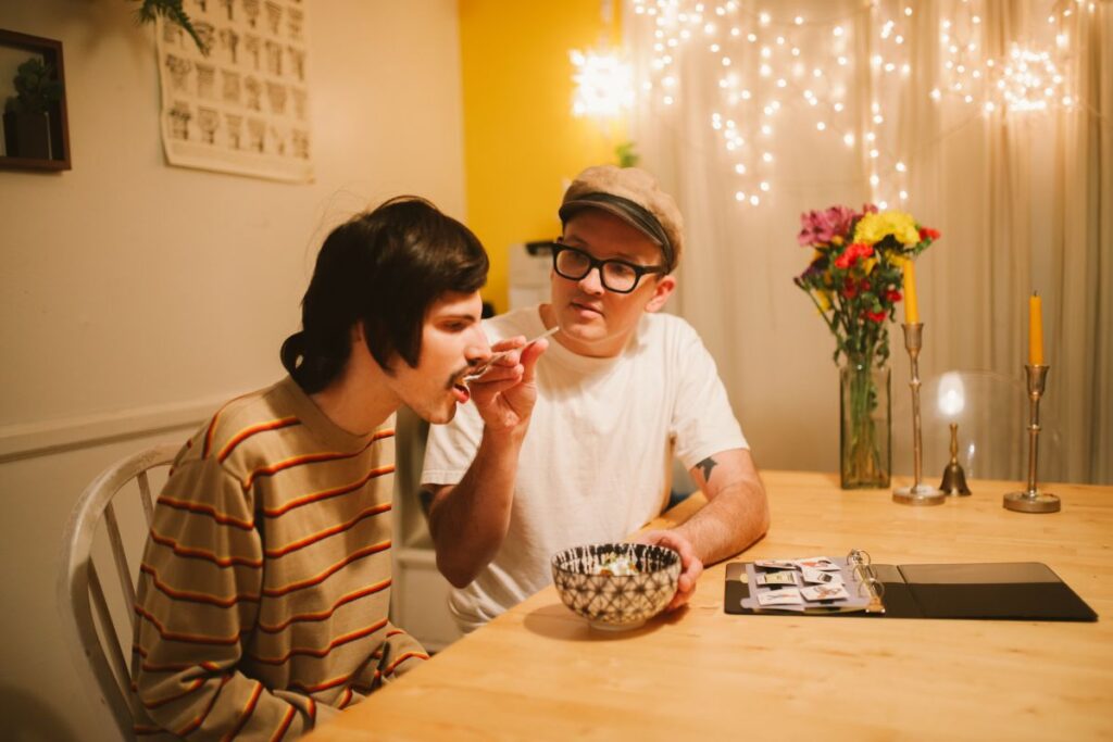 A man helps feed a young adult at home, highlighting the caregiving role of siblings of children with special needs.